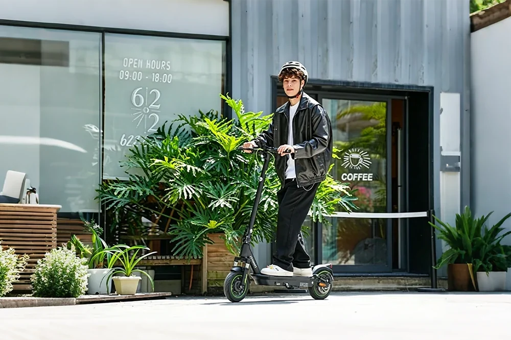 Young man wearing leather jacket and helmet riding portable electric scooter in front of cafe storefront with green plants, urban lifestyle e-scooter