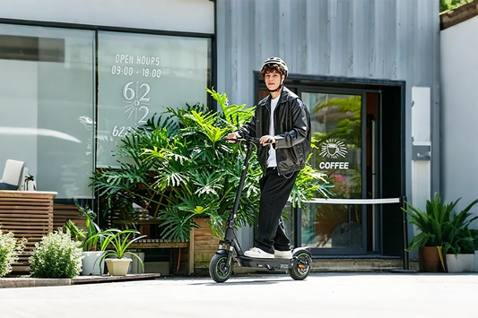 Young man wearing leather jacket and helmet riding portable electric scooter in front of cafe storefront with green plants, urban lifestyle e-scooter