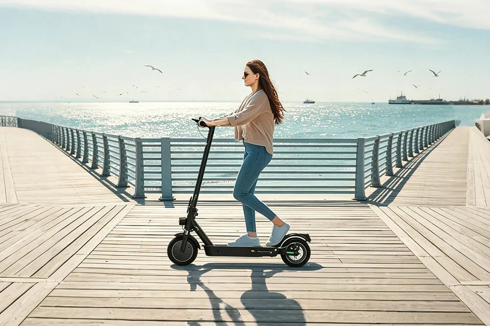 Foldable electric scooter being ridden by woman on wooden seaside pier with ocean backdrop